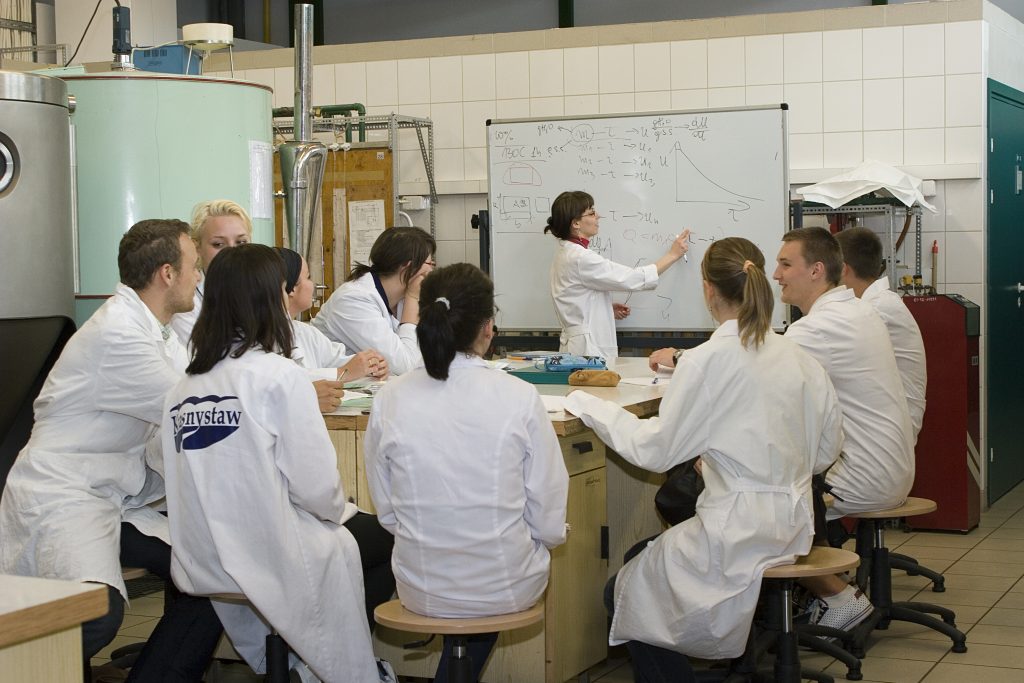 Grupa studentów i studentek ubranych w fartuchy laboratoryjne znajduje się na hali półtechniki w czasie zajęć. Siedzą przy dużym stole, w tle widać rózne urządzenia i prowadzącą zajęcia piszącą na tablicy.