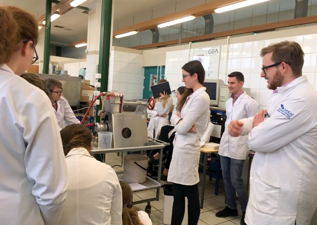 Grupa 7 studentów i studentek ubranych w fartuchy laboratoryjne znajduje się na hali półtechniki w czasie zajęć. Obsługują prasę filtracyjną, obok stoi prowadzący zajęcia, wszyscy się uśmiechają
