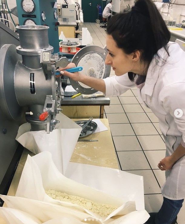 Studentka ubrana w fartuch laboratoryjny znajduje się na hali półtechniki. schylona nad otwartym z boku urządzeniem wygarnia z niego pędzelkiem proszek poddany aglomeracji. na stole obok leżą na arkuszach bibuł inne proszki. w tle widać inne urządzenia i innego studenta w fartuchu