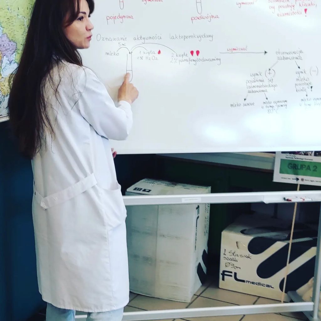 Widoczna jest biała szkolna tablica, na której jest narysowany schemat wykonania oznaczenia aktywności enzymu. Stoi przy niej naukowczyni ubrana w fartuch laboratoryjny, pokazuje palcem fragment schematu patrząc na kogoś słuchającego niewidocznego na zdjęciu