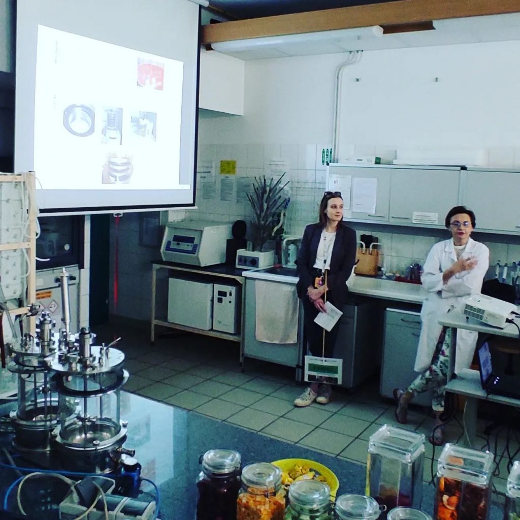 Widiczne znętrze laboratorium, słoiki z suszoną żywnością, sprzęt laboratoryjny na stołach. Na ekranie wyświetlona jest prezentacja o suszeniu, jedna naukowczyni tłumaczy, druga stoi obok trzymajać tabliczkę z nazwą wydziału
