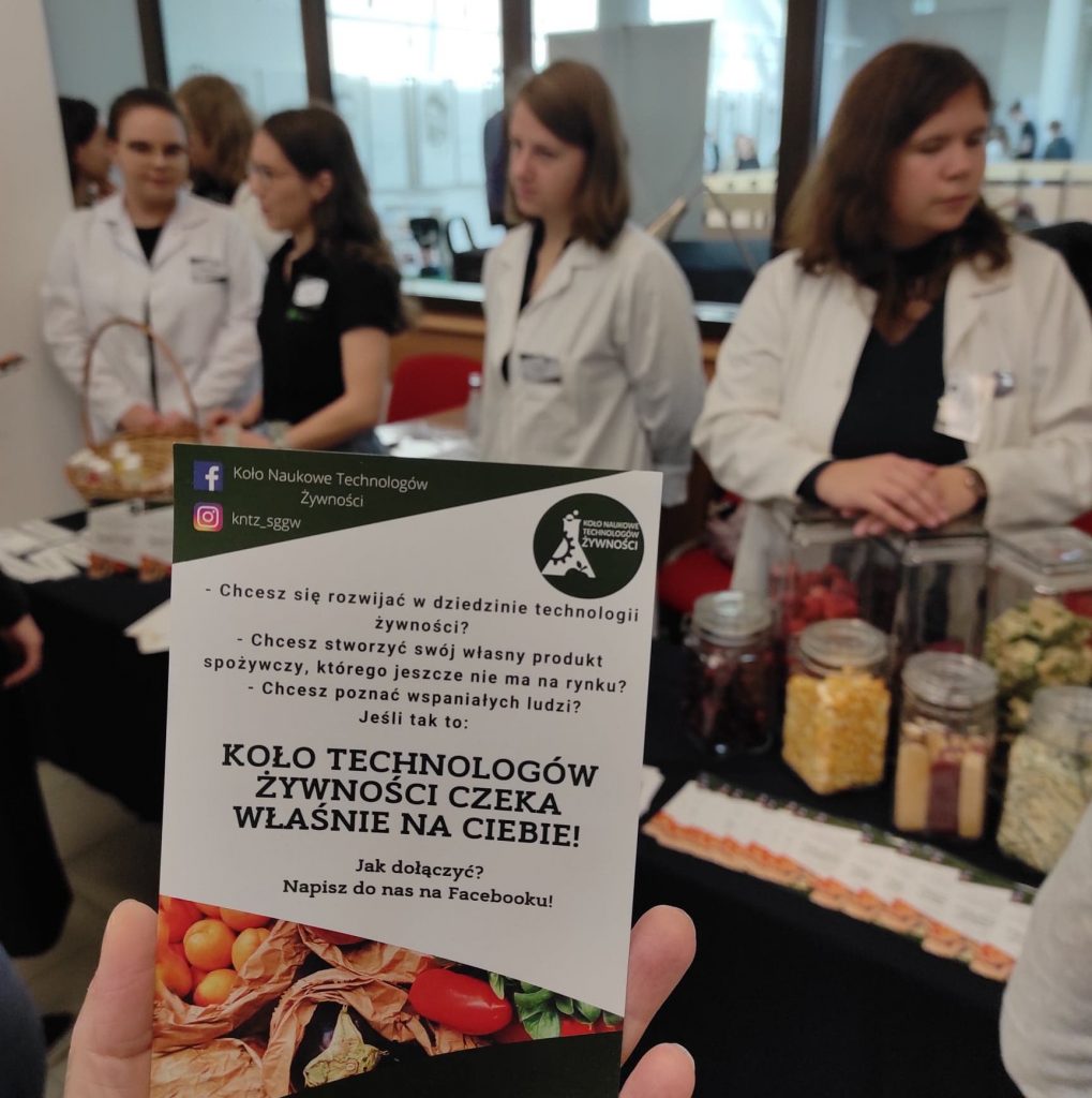 Zdjęcie ulotki Koła Naukowego Technologów Żywności zrobione na tle grupy 4 studentów. Studenci stoją prezentując zgromadzonej publiczności słoiki z suszoną żywnością i tłumacząc czym zajmuje się ich koło naukowe