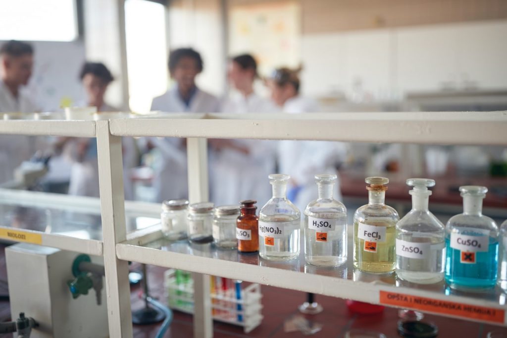 Butelki szklane ze szlifem wypełnione roztworami kwasów stoją na półce w laboratorium, w tle nieostro widoczna grupa studentów w fartuchach laboratoryjnych