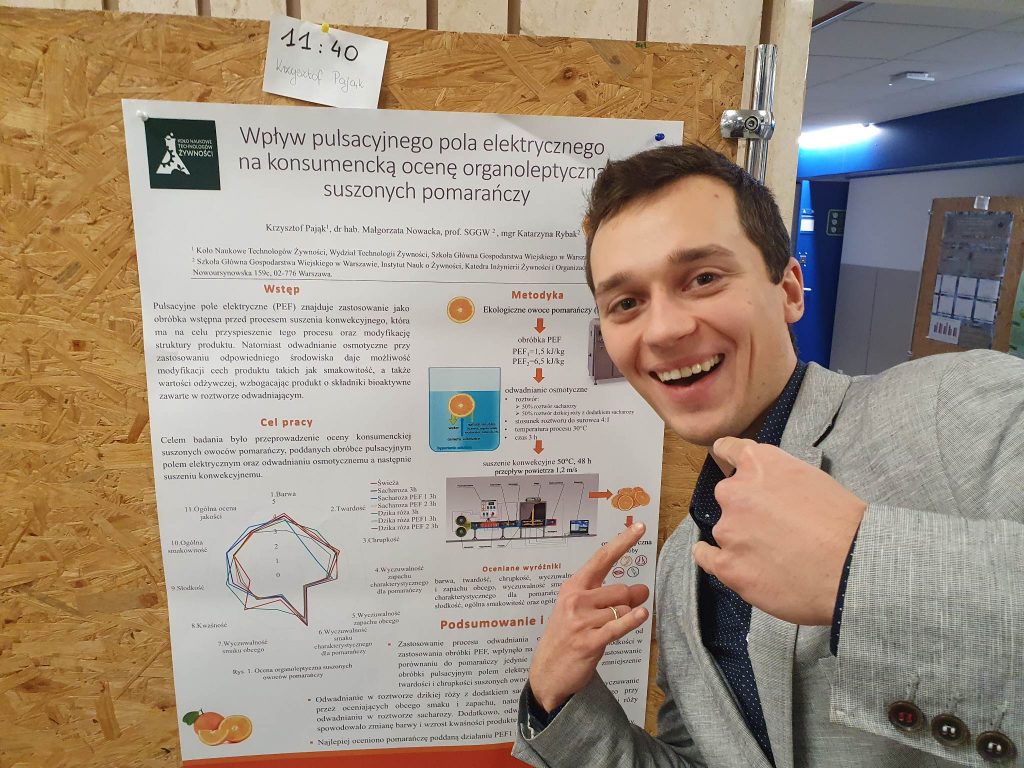 Widoczna jest tablica z przymocowanym na niej plakatem naukowym, Stoi przy niej uśmiechnięty student ubrany w marynarkę, wykonujący dłońmi gesty OK