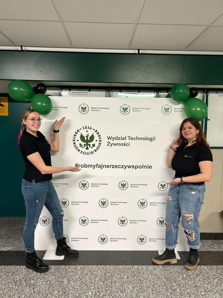Dwie studentki ubrane w czarne koszulki z napisem Wydział Technologii Żywności i jeansy stoją przed tablicą z nazwą wydziału i napisem róbmy fajne rzeczy wspólnie. Wskazują uśmiechnięte na ten napis