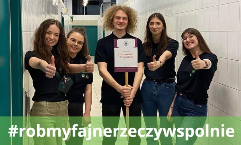Grupa 5 uśmiechniętych studentów i studentek stojących w korytarzu budynku wydziału. Osoby ubrane są w jednolite czarne koszulki z logo wydziału, mają podniesione kciuki w geście OK, jeden ze studentów trzyma tabliczkę z nazwą wydziału