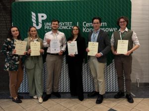 Grupa 6 studentów i studentek stoi w centrum konferencyjnym na tle ścianki z napisem Uniwersytet Przyrodniczy w Lublinie. Są elegancko ubrani, na szyi mają smycze z plakietkami i trzymają dyplomy poświadczające nagrodzenie ich wystapień na konferencji