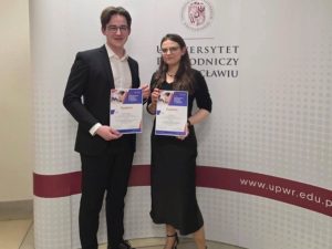 Student i studentka stoją na tle ścianki z napisem Uniwersytet Przyrodniczy we Wrocławiu, trzymają dyplomy przyznane za ich prezentacje na konferencji