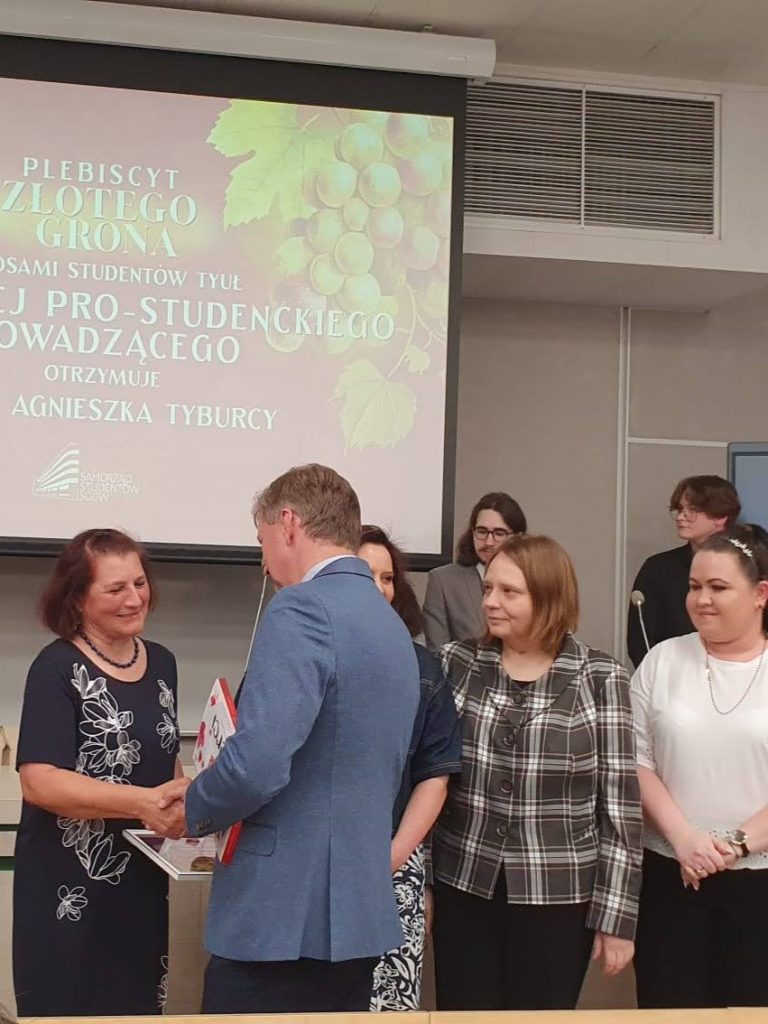 Prorektor wręcza naukowczyni nagrodę dla najbardziej pro-studenkiego prowadzącego przyznaną w plebiscycie samorządu studentów. 3 osoby stoją usmiechnięte obok, w tle ekran z wyswietloną nazwą nagrody