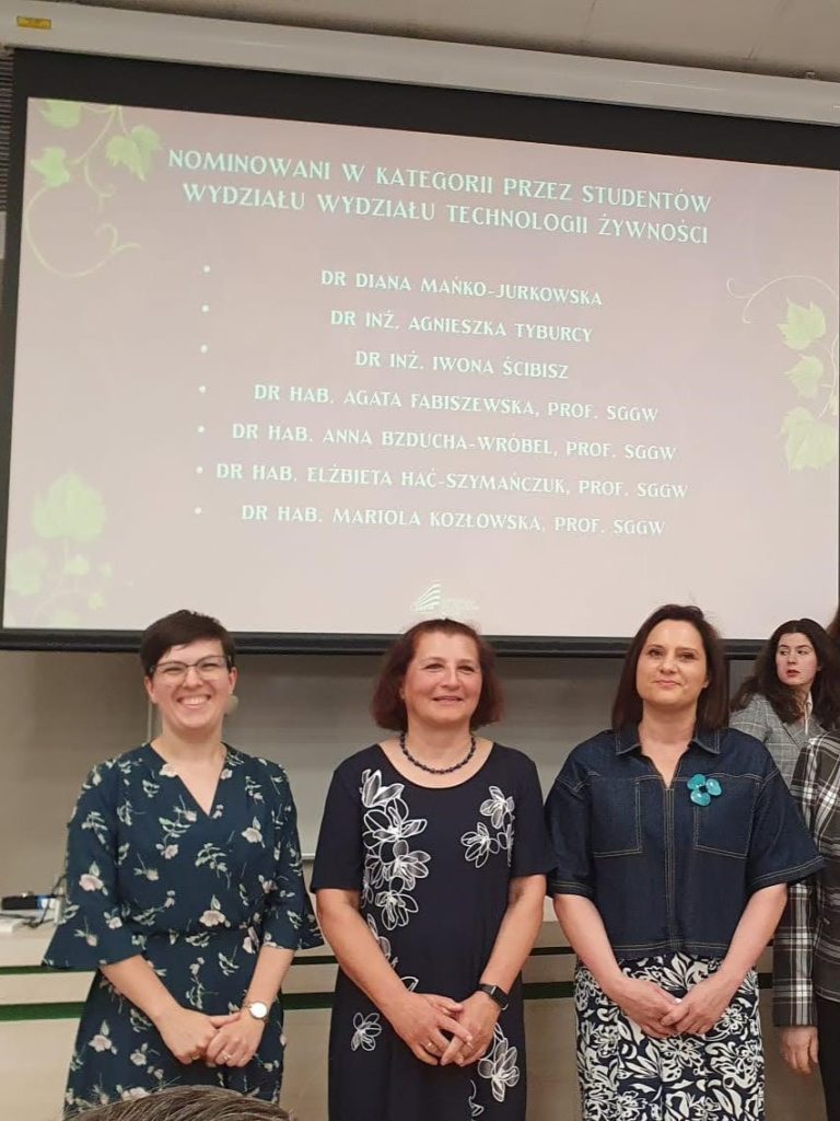 Grupa 3 elegancko ubranych naukowczyń stoi w auli w oczekiwaniu na ogłoszenie wyników plebiscytu. W tle ekran z nazwiskami