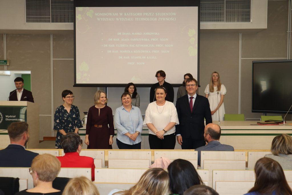 Grupa elegancko ubranych 5 naukowców i naukowczyń stoi w auli w oczekiwaniu na ogłoszenie wyników plebiscytu. W tle ekran z nazwiskami i osoby prowadzące wydarzenie, z przodu widać kilka rzedów widowni