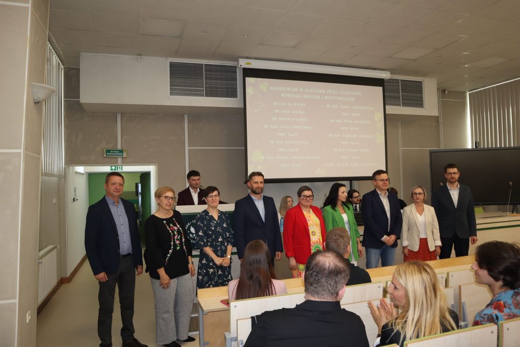 Grupa 9 elegancko ubranych naukowców i naukowczyń stoi w auli w oczekiwaniu na ogłoszenie wyniki plebiscytu. Widoczna od tyłu część widowni na auli, a na ekranie z tyłu lista nominowanych