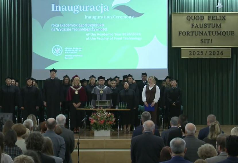 Widik na aulę, w której na podwyższeniu nauczyciele akademiccy wydziału stoją ubrani w uroczyste togi. Widoczna od tyłu stojąca część widowni