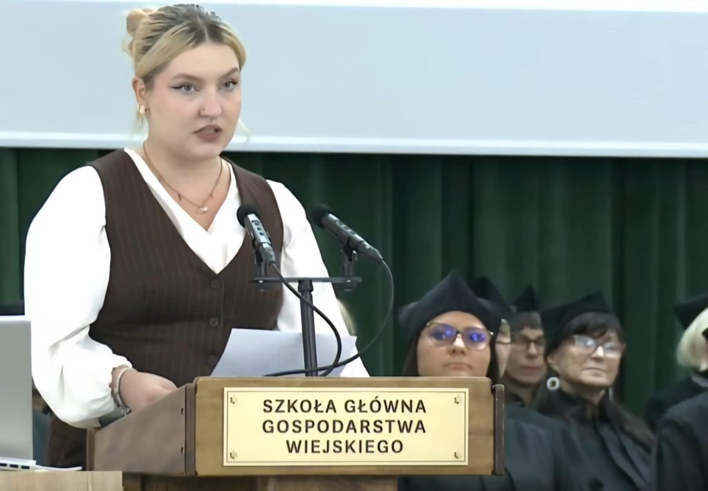 Elegancko ubrana studentka stoi przy mównicy w auli podczas inauguracji roku i wygłasza swoje przemówienie do nowych studentów. W tle siedzi kadra wydziału w uroczystych togach